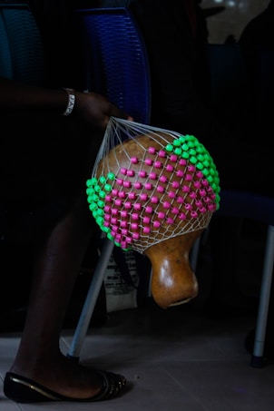 A hand is holding a traditional gourd shaker adorned with a colorful net made of pink and green beads. The background includes chairs, and the scene appears to capture a moment in a dark, indoor setting.