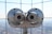 A pair of large silver binoculars mounted on a platform with a metal grid backdrop. The focus is on the metallic surface with visible wear and tear, and the background is softly blurred.