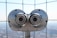 A pair of large silver binoculars mounted on a platform with a metal grid backdrop. The focus is on the metallic surface with visible wear and tear, and the background is softly blurred.