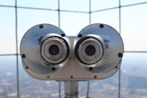 A pair of large silver binoculars mounted on a platform with a metal grid backdrop. The focus is on the metallic surface with visible wear and tear, and the background is softly blurred.