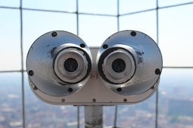 A pair of large silver binoculars mounted on a platform with a metal grid backdrop. The focus is on the metallic surface with visible wear and tear, and the background is softly blurred.
