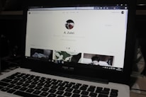 A laptop displaying a social media profile page with various elements such as a profile picture, username, and links. Below the main header, there are sections for photos and collections, showing preview images. The laptop keyboard is visible in the lower part of the image.