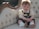 A baby with light brown hair is sitting on a vintage cream-colored couch with decorative wood accents. The baby is wearing a cream shirt with a green bowtie and green suspenders, along with white socks and brown shoes. The baby has a cheerful expression with a big smile.