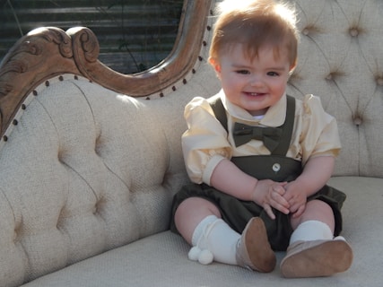 A baby with light brown hair is sitting on a vintage cream-colored couch with decorative wood accents. The baby is wearing a cream shirt with a green bowtie and green suspenders, along with white socks and brown shoes. The baby has a cheerful expression with a big smile.