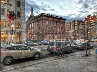 A city street scene featuring historic buildings and modern architectural styles. Cars are parked along the street, and bicycles are visible nearby. A decorated wreath hangs from a lamppost, suggesting a festive holiday season. The sky is cloudy, adding a serene atmosphere to the urban environment.