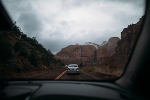 A scenic road trip view through a car windshield, featuring a highway flanked by towering reddish-brown cliffs on a cloudy day. The landscape includes scattered vegetation and a few vehicles ahead on the road. Snow caps the top of the cliffs in the distance, adding to the rugged natural setting.