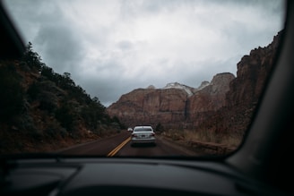 A scenic road trip view through a car windshield, featuring a highway flanked by towering reddish-brown cliffs on a cloudy day. The landscape includes scattered vegetation and a few vehicles ahead on the road. Snow caps the top of the cliffs in the distance, adding to the rugged natural setting.