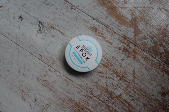 A round white container with blue and red text is placed on a worn wooden surface. The container is labeled 'Epok' with additional text in smaller font.