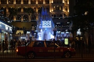 An illuminated historic building with a prominent 1881 Heritage sign in the center. The scene is bustling with people and features Christmas decorations, including small lights and trees. A red taxi is visible in the foreground, parked along the street.