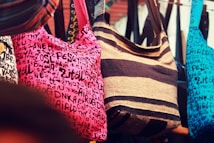 A selection of colorful fabric bags hanging closely together, each featuring distinct patterns and designs. The central bag has dark brown and tan stripes, while the others include a pink bag covered with black random letters and symbols and a similar design in blue. The bags are displayed in a market-like setting, creating a vibrant and eclectic visual array.
