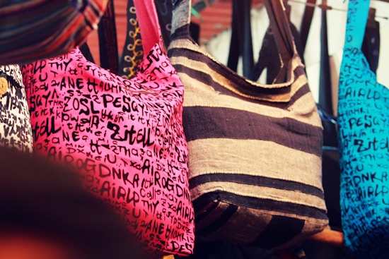 A selection of colorful fabric bags hanging closely together, each featuring distinct patterns and designs. The central bag has dark brown and tan stripes, while the others include a pink bag covered with black random letters and symbols and a similar design in blue. The bags are displayed in a market-like setting, creating a vibrant and eclectic visual array.