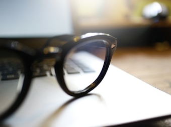 A pair of black-rimmed eyeglasses is resting on the closed lid of a laptop. The background is softly blurred, with warm lighting creating a cozy atmosphere.