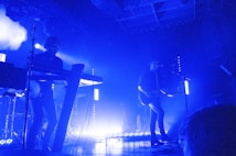 A dimly lit concert stage features two musicians. The scene is dominated by blue lighting, creating a moody atmosphere. One musician plays the keyboard while the other sings into a microphone and plays an electric guitar. Bright stage lights frame the performance area, casting shadows and emphasizing the silhouettes of the performers.