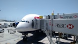 An airplane is parked at an airport gate, connected to a jet bridge marked with the number 150. The HSBC logo is prominently displayed on the jet bridge. Ground service vehicles and equipment are visible on the tarmac, with personnel attending to the aircraft. The sky is clear and blue, indicating good weather conditions.