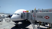 An airplane is parked at an airport gate, connected to a jet bridge marked with the number 150. The HSBC logo is prominently displayed on the jet bridge. Ground service vehicles and equipment are visible on the tarmac, with personnel attending to the aircraft. The sky is clear and blue, indicating good weather conditions.