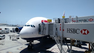 An airplane is parked at an airport gate, connected to a jet bridge marked with the number 150. The HSBC logo is prominently displayed on the jet bridge. Ground service vehicles and equipment are visible on the tarmac, with personnel attending to the aircraft. The sky is clear and blue, indicating good weather conditions.