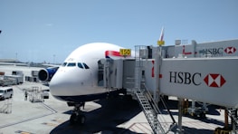 An airplane is parked at an airport gate, connected to a jet bridge marked with the number 150. The HSBC logo is prominently displayed on the jet bridge. Ground service vehicles and equipment are visible on the tarmac, with personnel attending to the aircraft. The sky is clear and blue, indicating good weather conditions.