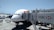 An airplane is parked at an airport gate, connected to a jet bridge marked with the number 150. The HSBC logo is prominently displayed on the jet bridge. Ground service vehicles and equipment are visible on the tarmac, with personnel attending to the aircraft. The sky is clear and blue, indicating good weather conditions.