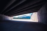 A structured concrete passage with a series of beams is framing a clear view of a distant mountain range. Sunlight illuminates a section of the concrete floor leading to the vibrant landscape of green trees and blue sky.