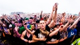 A large crowd of people at a music festival, with many individuals raising their hands enthusiastically. The image captures the excitement and energy of the audience, with people tightly packed together near the front barrier. The attendees display casual clothing and accessories typical of an outdoor event.