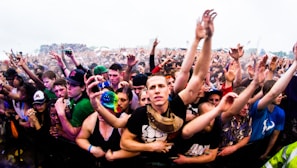 A large crowd of people at a music festival, with many individuals raising their hands enthusiastically. The image captures the excitement and energy of the audience, with people tightly packed together near the front barrier. The attendees display casual clothing and accessories typical of an outdoor event.