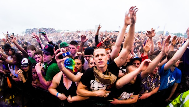 A large crowd of people at a music festival, with many individuals raising their hands enthusiastically. The image captures the excitement and energy of the audience, with people tightly packed together near the front barrier. The attendees display casual clothing and accessories typical of an outdoor event.