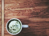 A white cup filled with a frothy green beverage, possibly matcha latte, sits on a wooden table with a textured grain pattern.