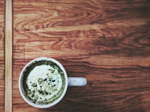 A white cup filled with a frothy green beverage, possibly matcha latte, sits on a wooden table with a textured grain pattern.
