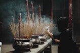 A person is placing incense sticks in a large ornate incense burner inside a dimly lit temple. Smoke rises from the burning incense, creating a mystical atmosphere. The burners are intricately decorated with carvings and contain a large number of incense sticks, some of which are quite long and red-tipped.
