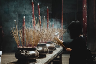 A person is placing incense sticks in a large ornate incense burner inside a dimly lit temple. Smoke rises from the burning incense, creating a mystical atmosphere. The burners are intricately decorated with carvings and contain a large number of incense sticks, some of which are quite long and red-tipped.