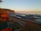 A picturesque view of a coastal area during what appears to be sunset or sunrise. The scene features a wooden house with a red roof situated near a rocky beach. The tide is low, revealing wet sand and various tidal patterns. A few people are walking or standing near the shore, admiring the landscape. In the background, a forested cliff extends into the distance, and the sky is bathed in warm hues.