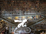 The image depicts an expansive, multi-story library filled with countless books on several shelves. A white sculpture of a flying figure on a bicycle is suspended in the center. People are browsing and walking around the library. Staircases connect different levels, and the overall setting appears bustling and dynamic.