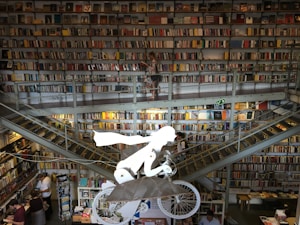 The image depicts an expansive, multi-story library filled with countless books on several shelves. A white sculpture of a flying figure on a bicycle is suspended in the center. People are browsing and walking around the library. Staircases connect different levels, and the overall setting appears bustling and dynamic.
