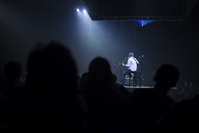 A person plays an acoustic guitar while seated on a stool on stage. Dim lighting creates a spotlight effect on the performer, while the rest of the stage and audience are in shadow. Audience members are silhouetted in the foreground.