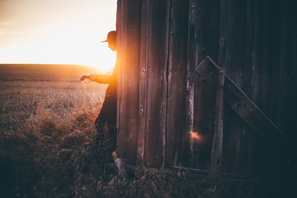 silhouette of man leaning on gray wooden house during sunset