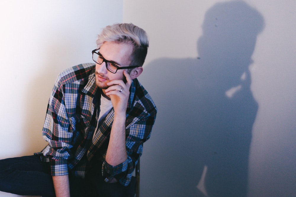 man wearing black, blue, and red plaid collared button-up long-sleeved shirt and eyeglasses sitting and putting left hand on his face