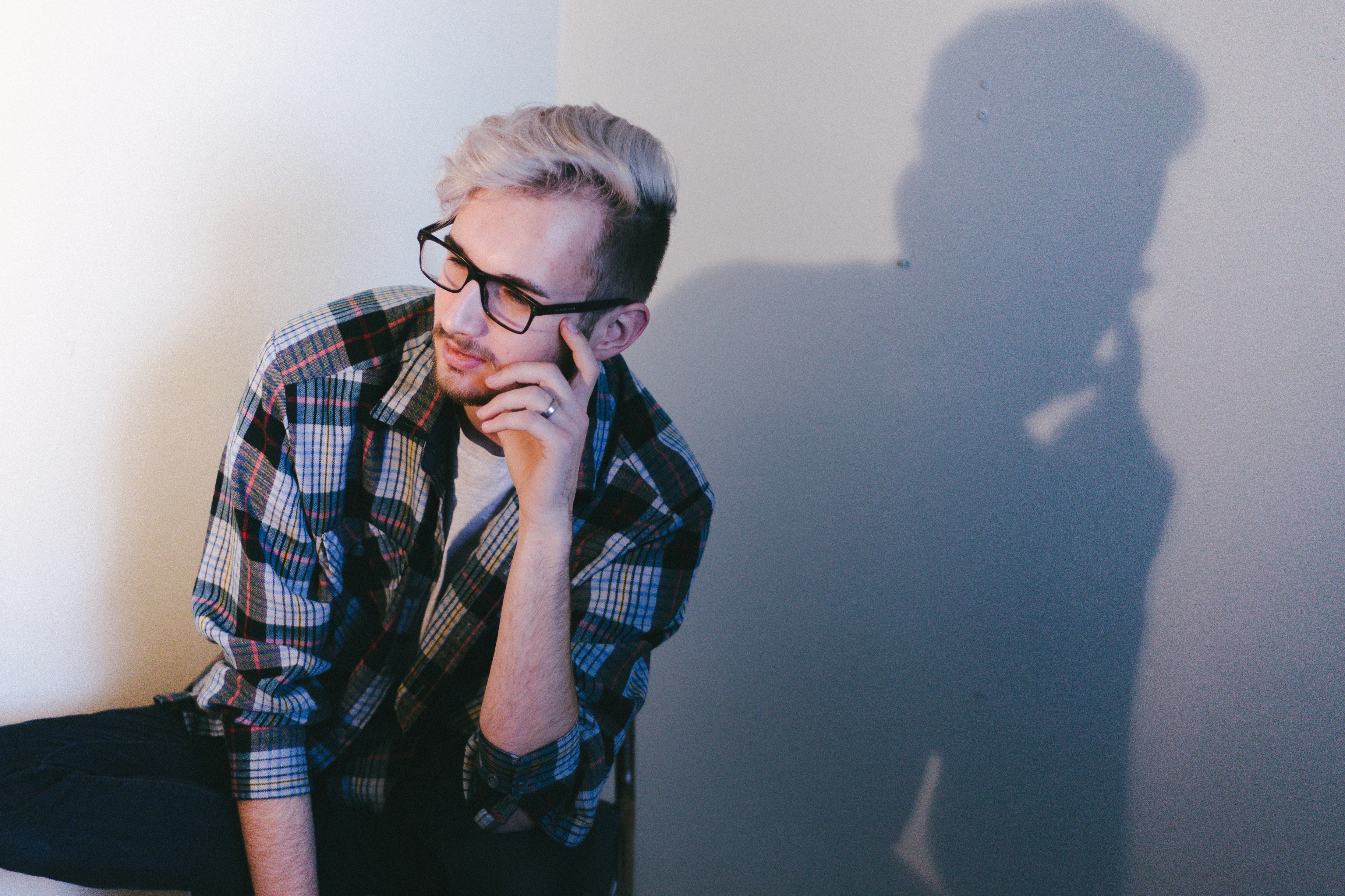 man wearing black, blue, and red plaid collared button-up long-sleeved shirt and eyeglasses sitting and putting left hand on his face