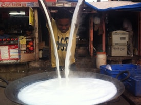 A man is pouring a large quantity of milk from two metal containers into a large cast iron pan. The milk is steaming as it flows in two streams, indicating it is hot. The setting appears to be a street-side food stall with colorful posters and a makeshift appearance. There are blue crates and some glassware visible nearby.