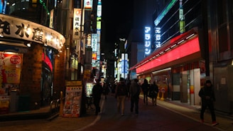 A vibrant urban street scene at night with numerous illuminated signs in various colors and languages. The street is bustling with people walking along the sidewalks beside shops and restaurants, creating a lively and energetic atmosphere.