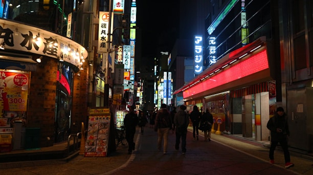 A vibrant urban street scene at night with numerous illuminated signs in various colors and languages. The street is bustling with people walking along the sidewalks beside shops and restaurants, creating a lively and energetic atmosphere.