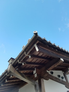 A traditional wooden roof structure features intricate wooden beams and tiles, set against a clear blue sky. The architecture highlights a harmonious blend of wood and ceramic materials.