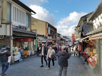 A bustling street scene in a traditional Japanese shopping area. Several people are walking and shopping, surrounded by buildings with Japanese signage and architecture. The sky is partly cloudy, and a sense of liveliness permeates the scene as people engage with vendors and each other.