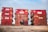 A person is sitting on the ground in front of three large red dumpsters, each with visible signs of wear, rust, and some graffiti. The scene is set outdoors under a cloudy sky, and the dumpsters appear to be aligned in a row. The person is sitting cross-legged, looking towards the camera.
