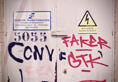 A metal door with a sign indicating it is a transformer station, accessible only to authorized personnel, is displayed. The door also has a warning sign about high voltage and the danger of death. Various graffiti, including the numbers 5055 and the words 'Faker' and 'GTK' in both red and black paint, are visible on the surface.
