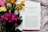 An open book displaying a chapter titled 'What Prevents You from Finding Love' lies on a white and pink patterned fabric. Next to the book is a vibrant bouquet of flowers including pink, yellow, and orange blooms with deep green leaves.