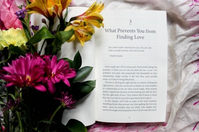 An open book displaying a chapter titled 'What Prevents You from Finding Love' lies on a white and pink patterned fabric. Next to the book is a vibrant bouquet of flowers including pink, yellow, and orange blooms with deep green leaves.