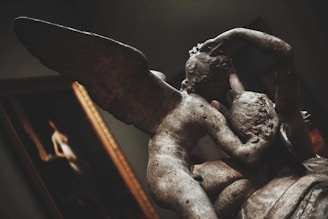 A close-up of a classical sculpture depicting two figures embracing. One figure has wings, suggesting it might represent an angel or a mythological character. The texture and shadows of the stone are highlighted, giving a sense of depth and antiquity. In the background, a framed painting is partially visible.