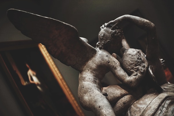 A close-up of a classical sculpture depicting two figures embracing. One figure has wings, suggesting it might represent an angel or a mythological character. The texture and shadows of the stone are highlighted, giving a sense of depth and antiquity. In the background, a framed painting is partially visible.