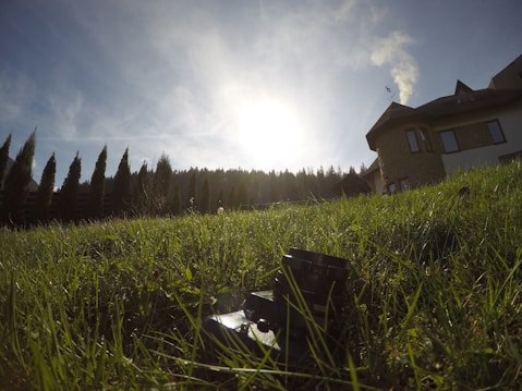 A wide-angle view from ground level showing a lush green lawn leading up to a house with a sloped roof in the background. The sky is clear and blue, with the sun shining brightly, casting long shadows. A line of tall trees borders the property, creating a serene and peaceful atmosphere.