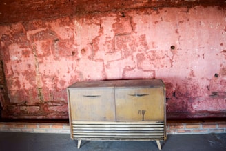 A vintage wooden cabinet with two doors is placed against a textured wall with a faded reddish hue. The wall, showing signs of wear, has a rough brick pattern and sporadic holes. The cabinet's surface is slightly worn, contributing to a rustic atmosphere.